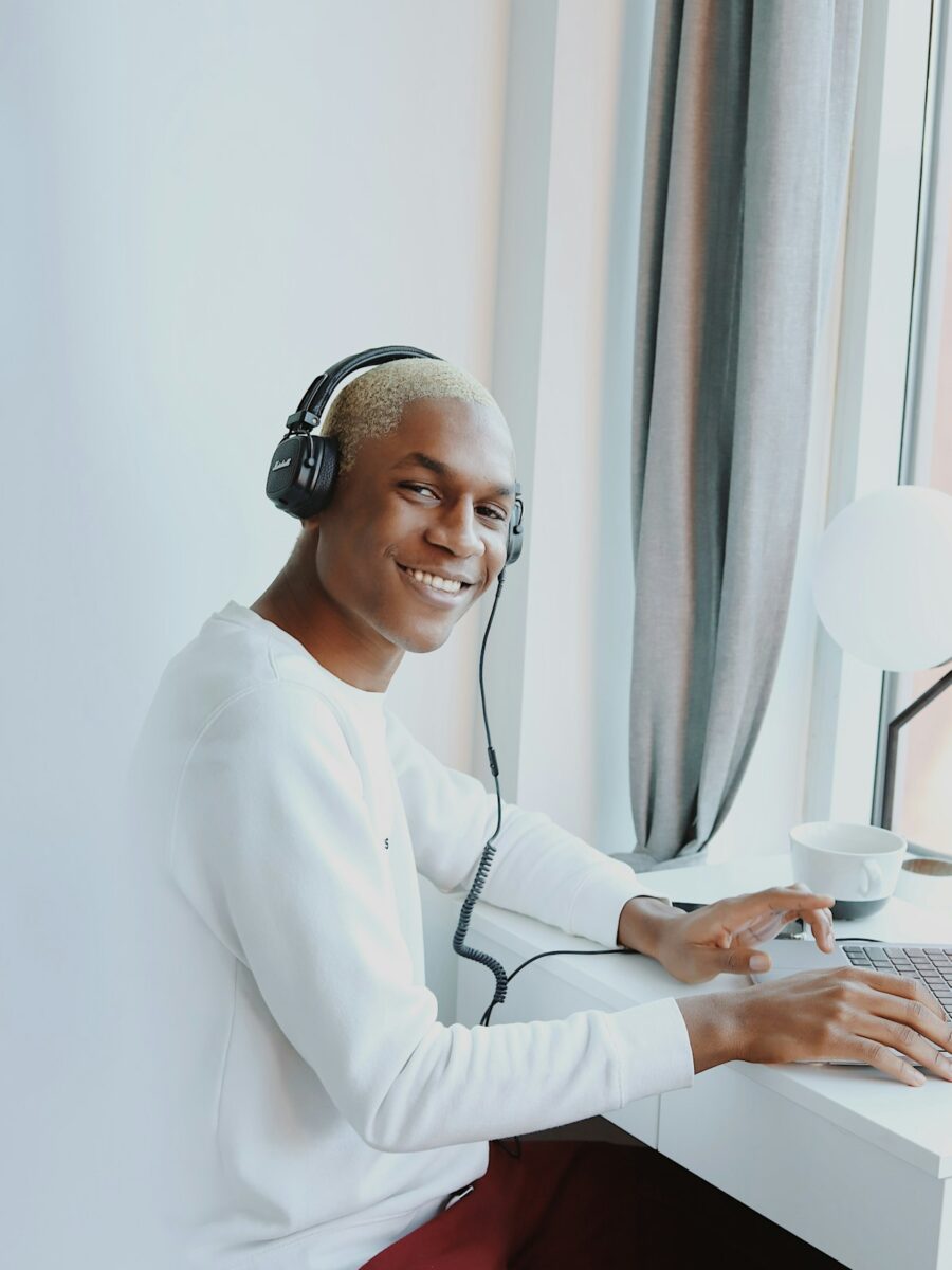 Een man met een koptelefoon op zit aan een bureau bij het raam en kijkt naar de camera