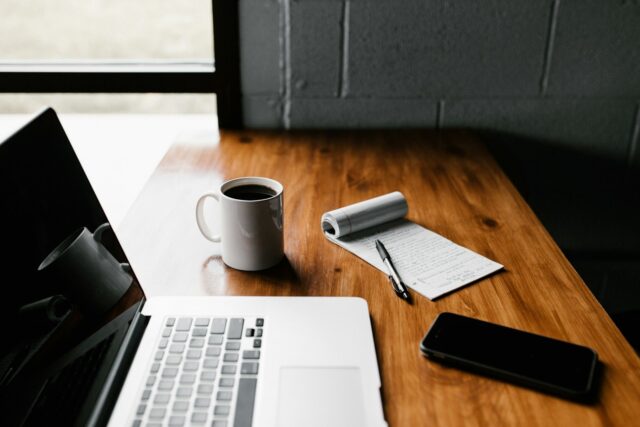 Een open laptop staat op een houten tafel naast een kop koffie en een notitieblok